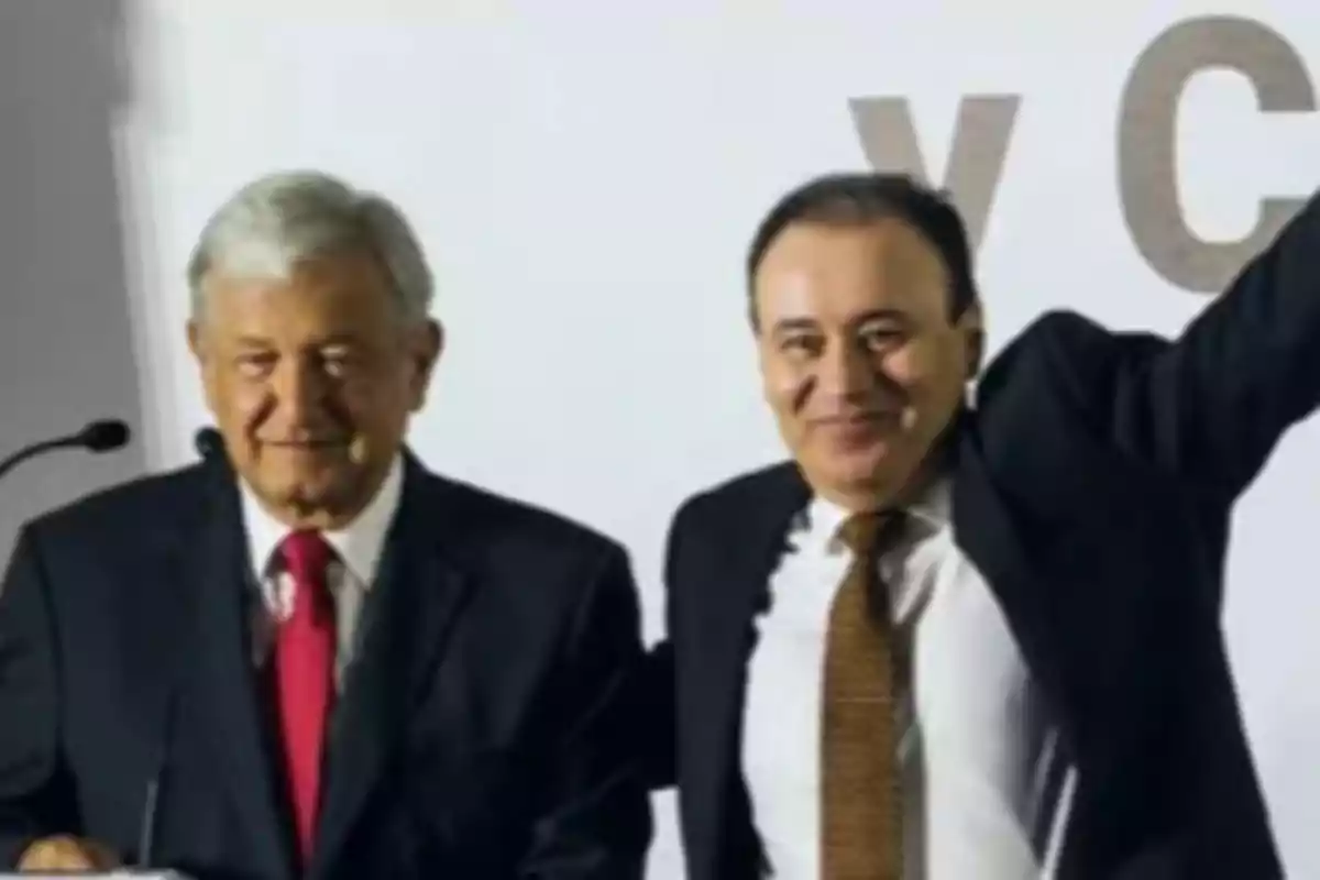 Dos hombres de traje oscuro y corbata posan juntos frente a un fondo claro, uno de ellos levanta el brazo.