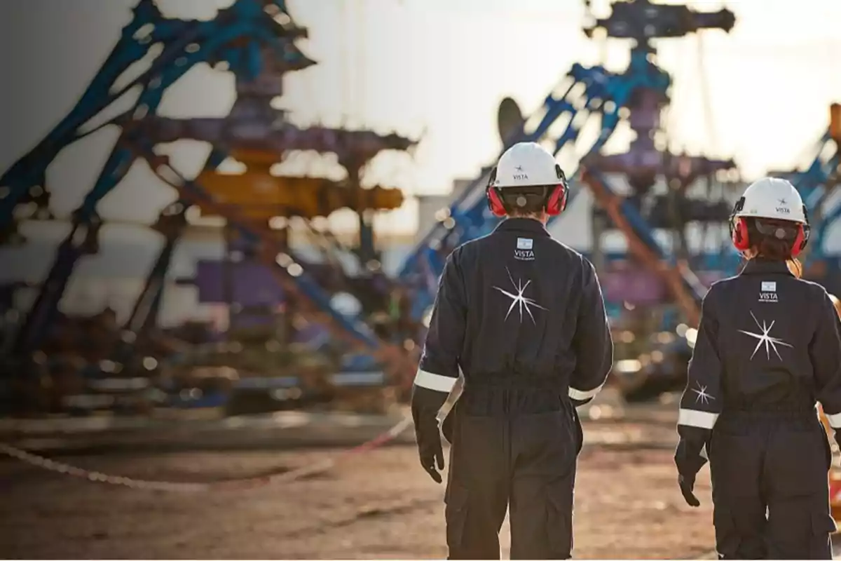 Dos personas con cascos y protectores auditivos caminan en un sitio industrial con maquinaria al fondo.