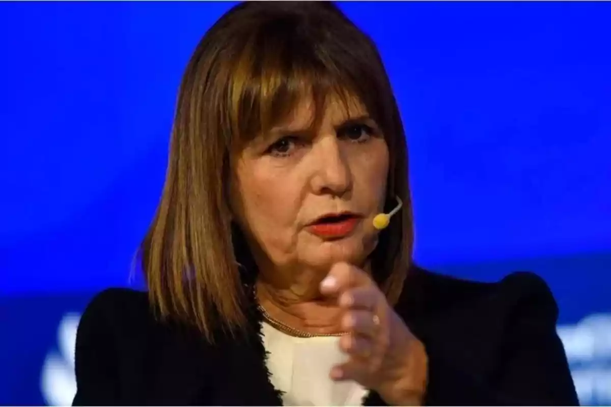 Una mujer hablando en un escenario con un micrófono de diadema y fondo azul.