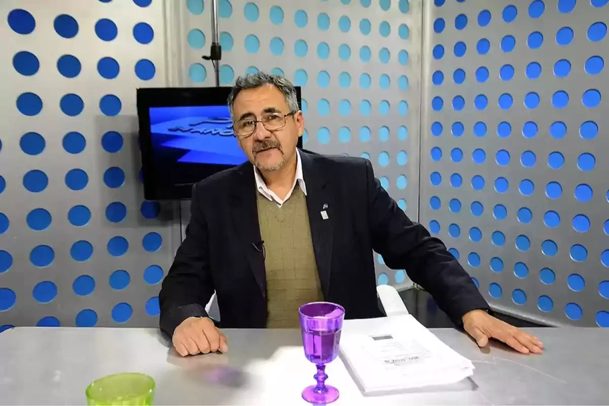 Middle-aged man with glasses and a beard sitting in a television studio with a background of perforated metal panels, a purple glass, and papers on the table. Middle-aged man with glasses and a beard sitting in a television studio with a background of perforated metal panels, a purple glass, and papers on the table.