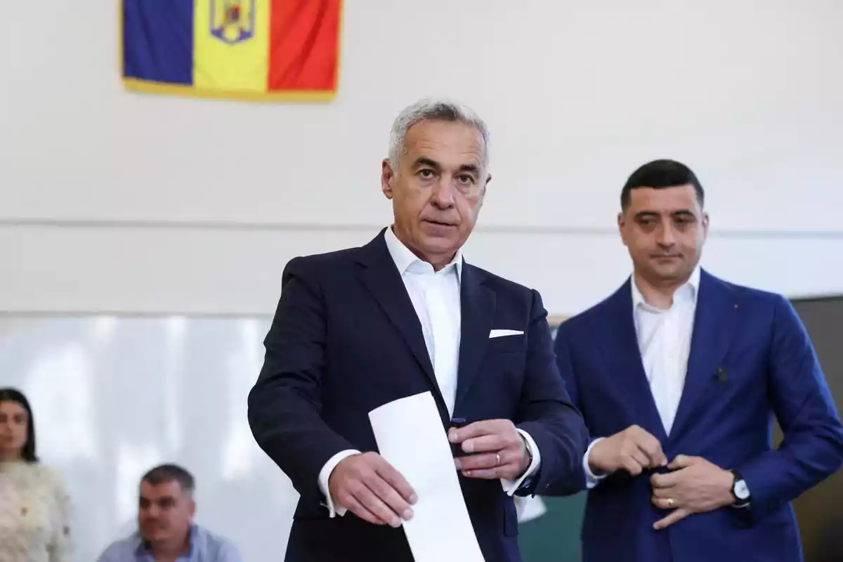 Gray-haired man in a dark suit casting a ballot into a box while another person in a blue suit watches him and a tricolor flag hangs on the wall in the background.