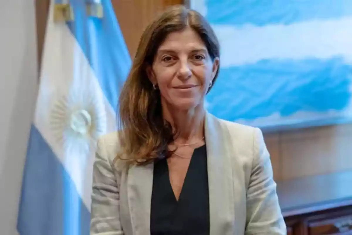 Una mujer de cabello castaño y chaqueta clara está de pie frente a una bandera argentina.