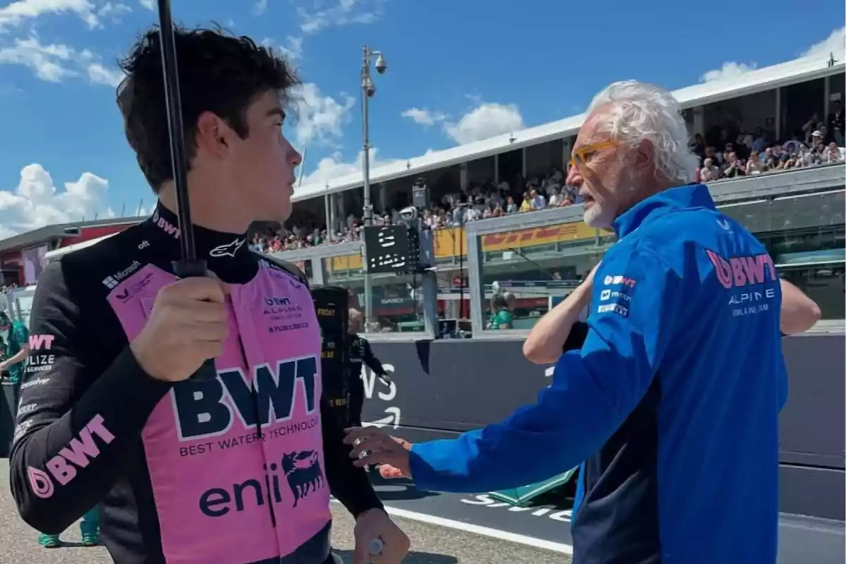Un piloto con uniforme rosa y negro sostiene un paraguas mientras conversa con un hombre de cabello canoso y gafas, ambos en una pista de carreras con público al fondo Un piloto con uniforme rosa y negro sostiene un paraguas mientras conversa con un hombre de cabello canoso y gafas, ambos en una pista de carreras con público al fondo