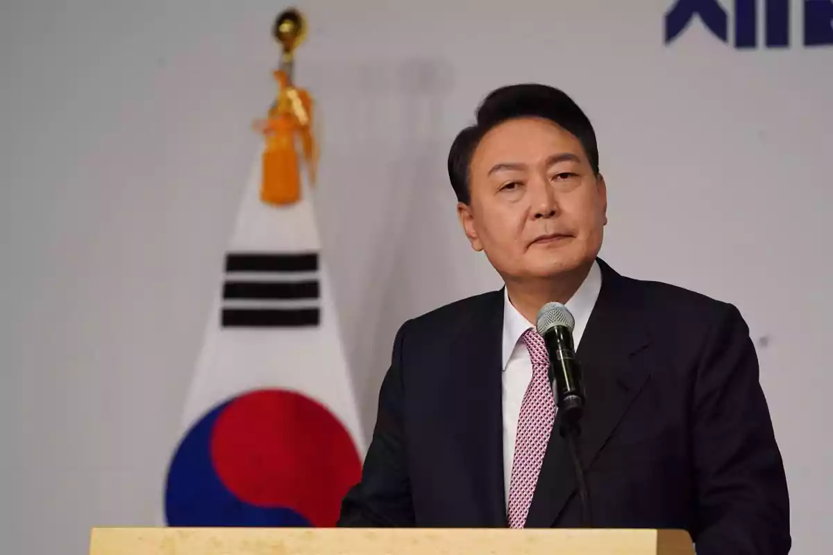 Hombre de traje oscuro hablando en un podio con un micrófono y una bandera de Corea del Sur al fondo Hombre de traje oscuro hablando en un podio con un micrófono y una bandera de Corea del Sur al fondo