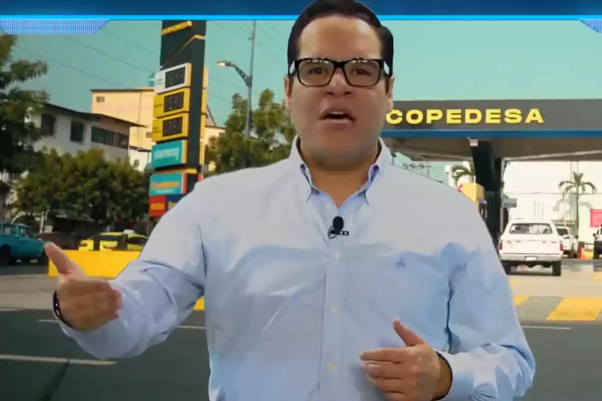 Un hombre con gafas y camisa azul está hablando frente a una estación de servicio con un cartel de precios visible al fondo.