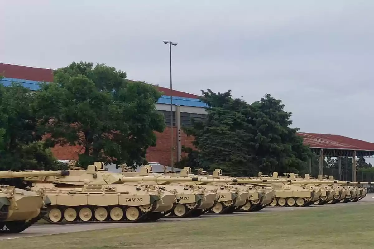 Fila de tanques TAM 2C estacionados en un área al aire libre junto a un edificio y árboles.