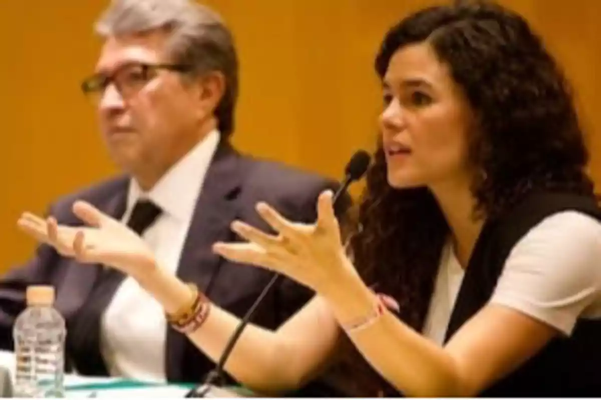 Two people sitting at a table during a conference, one of them speaking into a microphone and gesturing with their hands while the other listens alertly. Two people sitting at a table during a conference, one of them speaking into a microphone and gesturing with their hands while the other listens alertly.