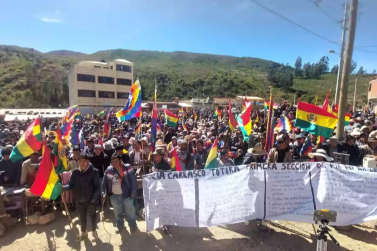 Una multitud de personas se reúne al aire libre sosteniendo banderas de Bolivia y wiphalas, mientras exhiben una pancarta grande con texto manuscrito, en un entorno rural con colinas y edificios al fondo. Una multitud de personas se reúne al aire libre sosteniendo banderas de Bolivia y wiphalas, mientras exhiben una pancarta grande con texto manuscrito, en un entorno rural con colinas y edificios al fondo.