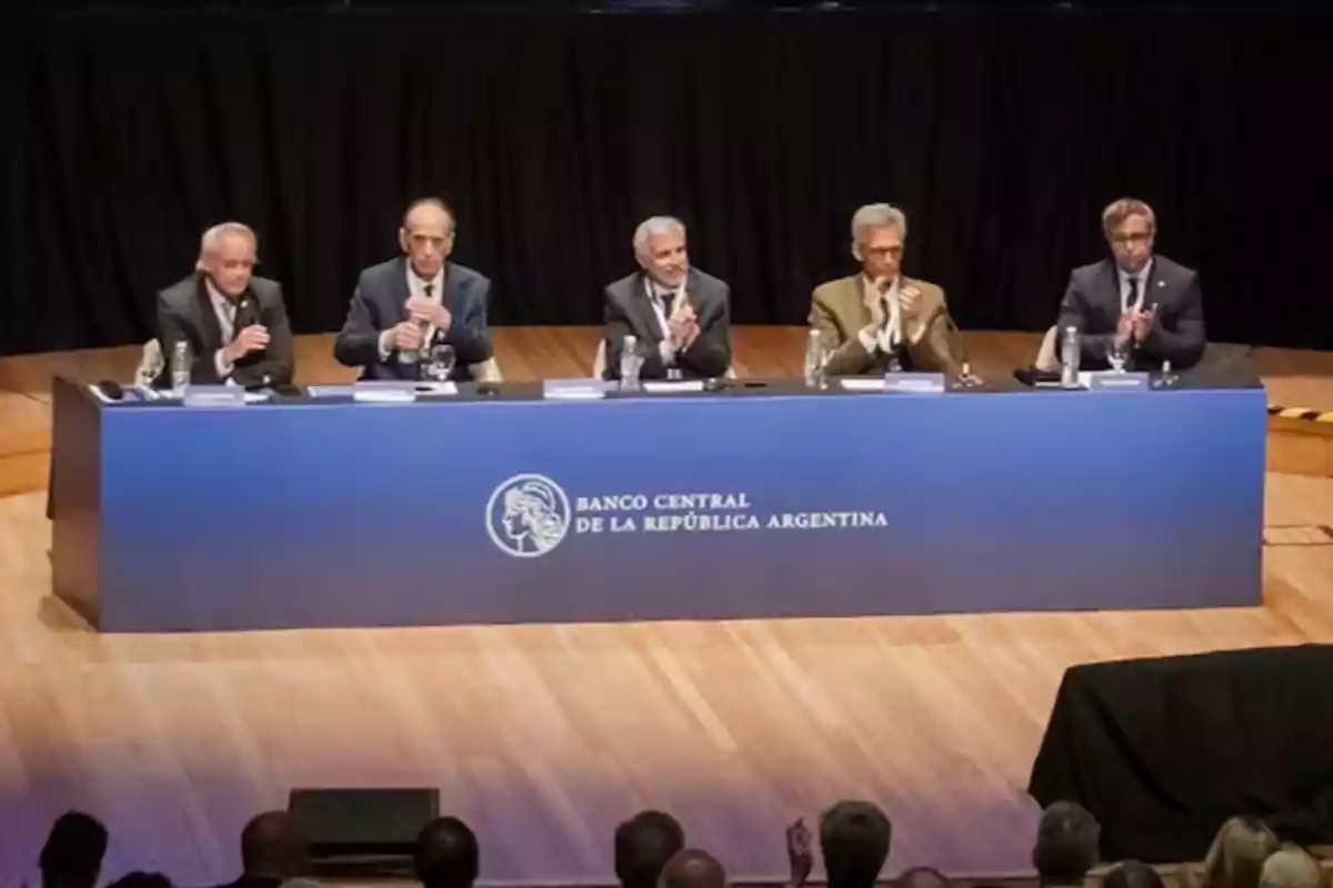 Cinco personas sentadas en una mesa azul del Banco Central de la República Argentina en un escenario de madera, aplaudiendo frente a una audiencia.