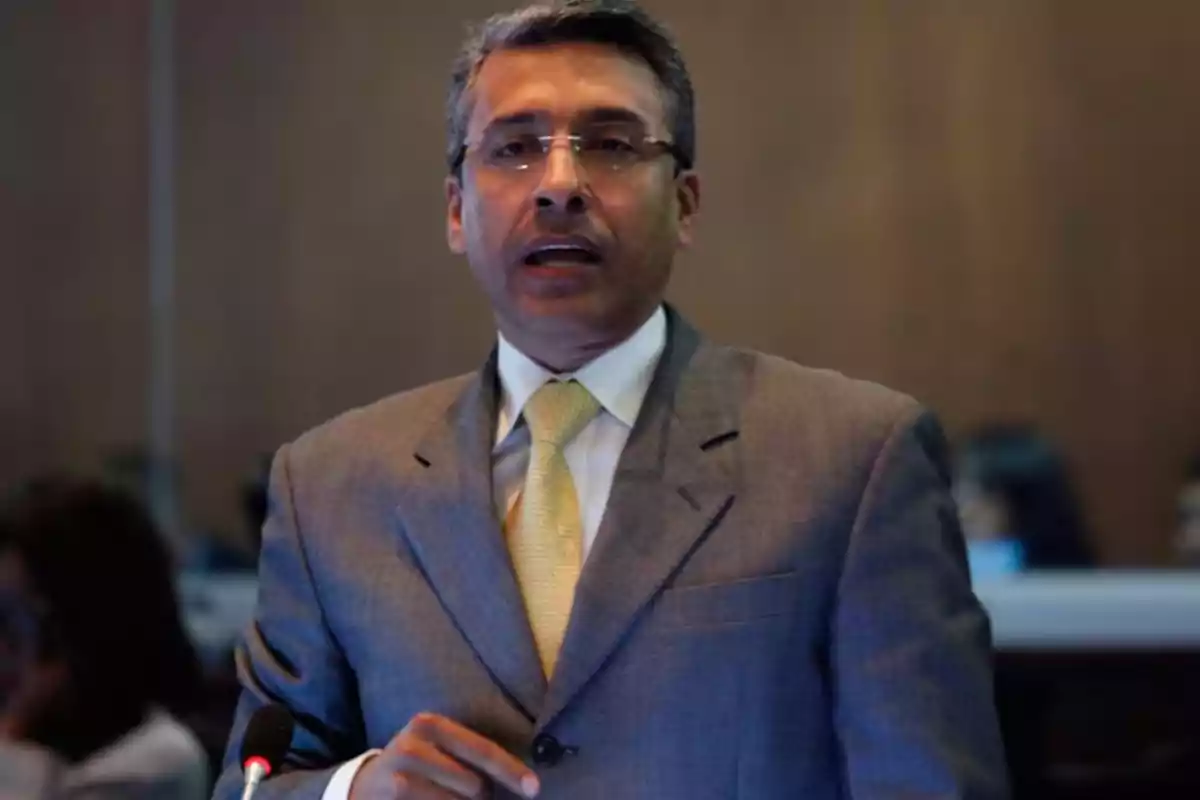 A man in a gray suit and yellow tie speaking into a microphone in a professional setting.