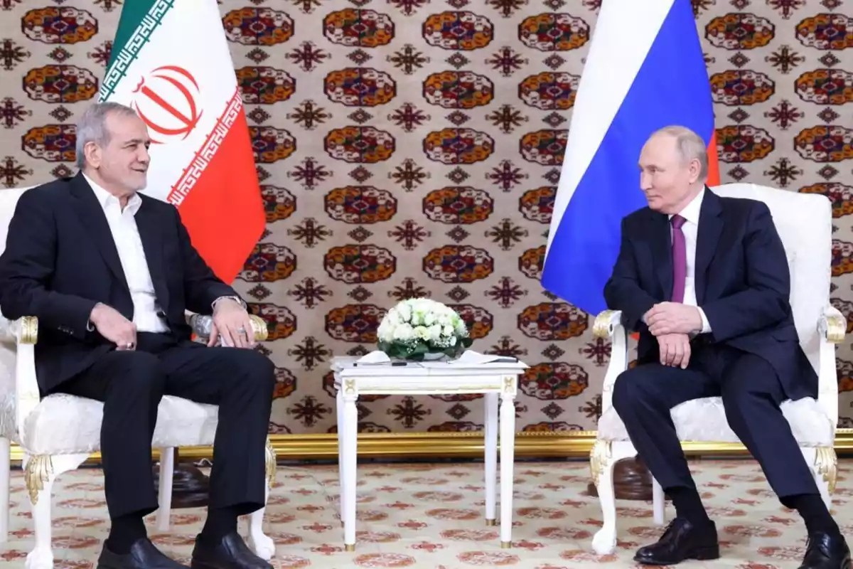 Dos hombres sentados en sillas blancas frente a frente con banderas de Irán y Rusia detrás de ellos y una mesa pequeña con flores en el centro.