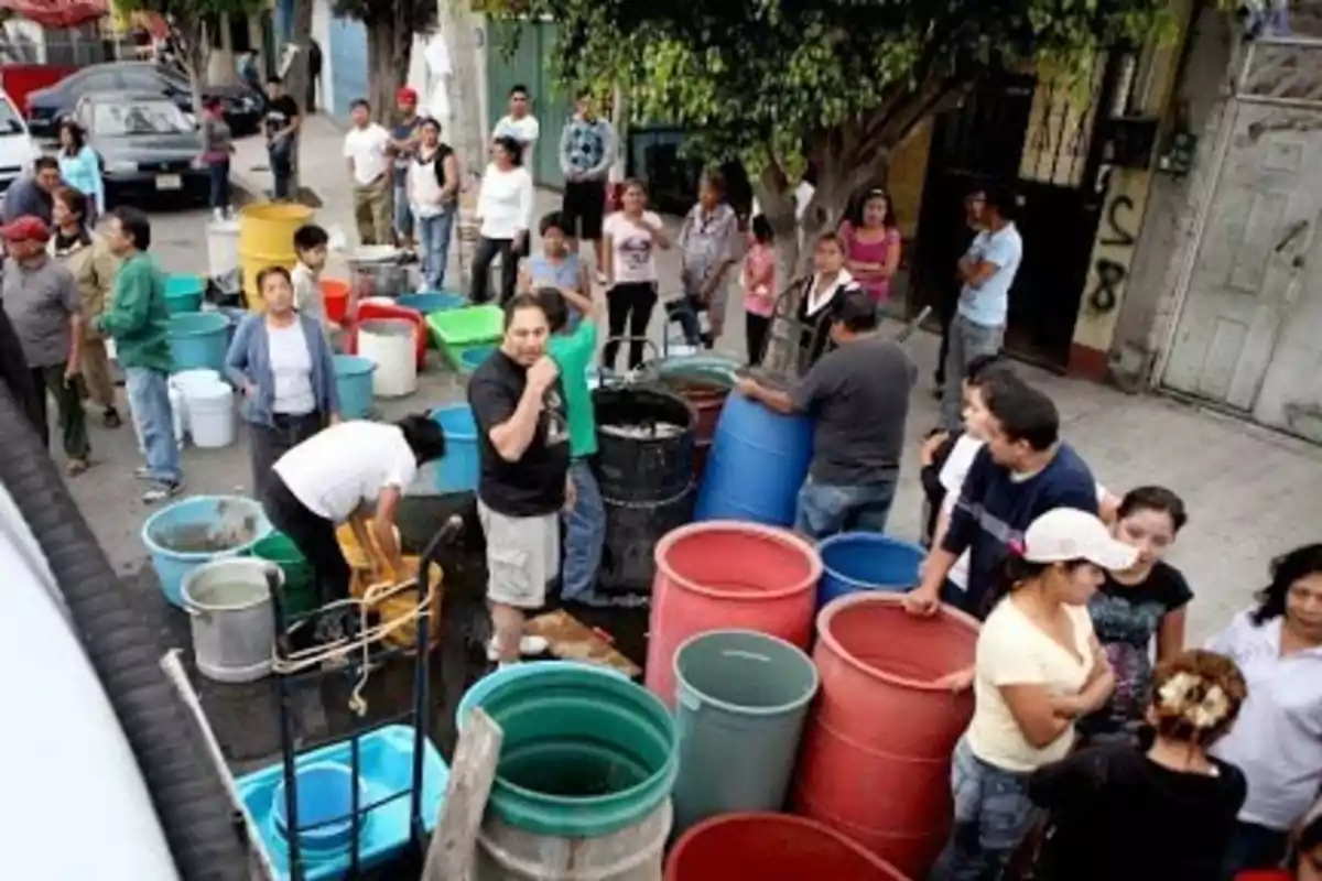 A group of people gathers in the street with several plastic containers of different colors, possibly to collect water.