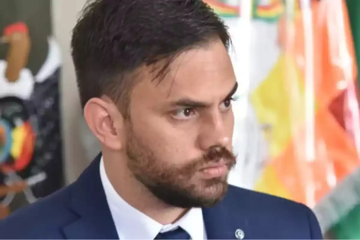 Hombre de cabello corto y barba con expresión seria vistiendo traje azul y corbata, fondo desenfocado con colores vivos Hombre de cabello corto y barba con expresión seria vistiendo traje azul y corbata, fondo desenfocado con colores vivos