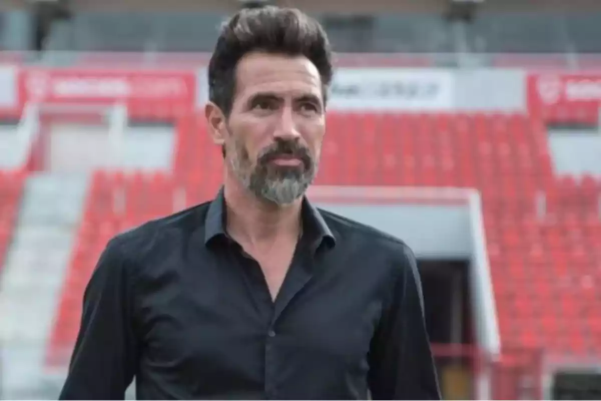 Hombre con barba y cabello oscuro en un estadio con asientos rojos de fondo.