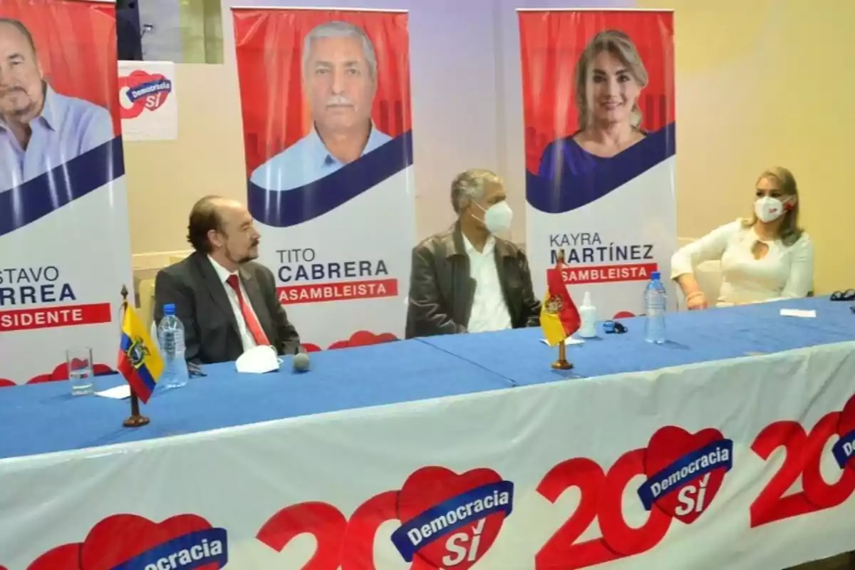 People sitting at a table with blue tablecloths and small flags, behind them are political campaign posters with images of candidates and the "Democracy Yes" logo.