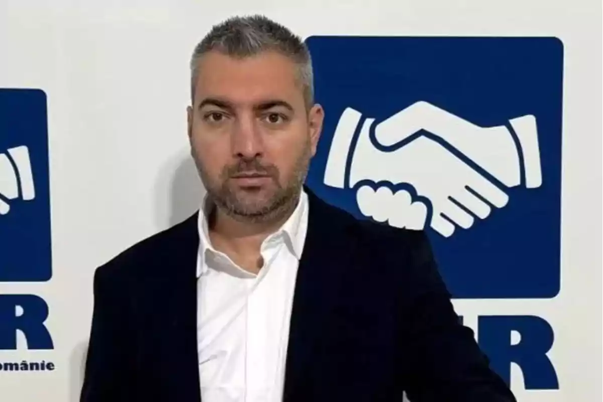 Un hombre de cabello corto y barba está de pie frente a un fondo con un logotipo de un apretón de manos. Un hombre de cabello corto y barba está de pie frente a un fondo con un logotipo de un apretón de manos.