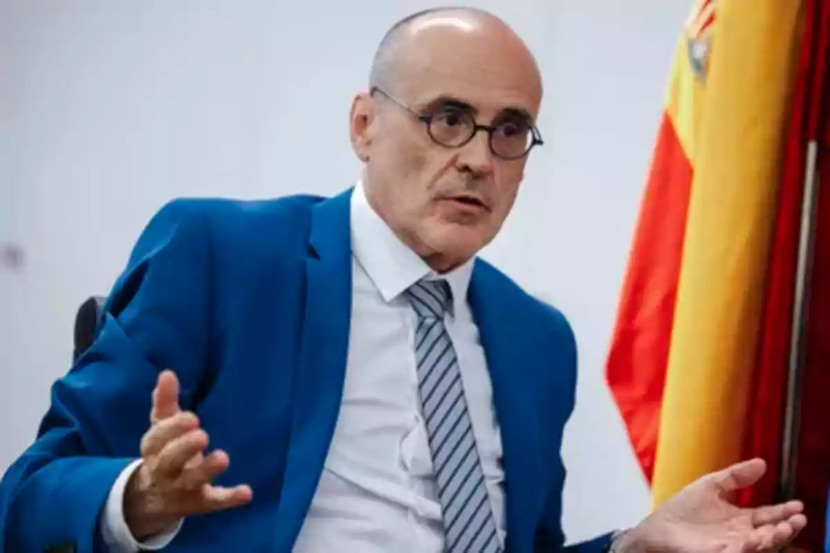 Hombre calvo con gafas y traje azul hablando gesticulando con una bandera española en el fondo.