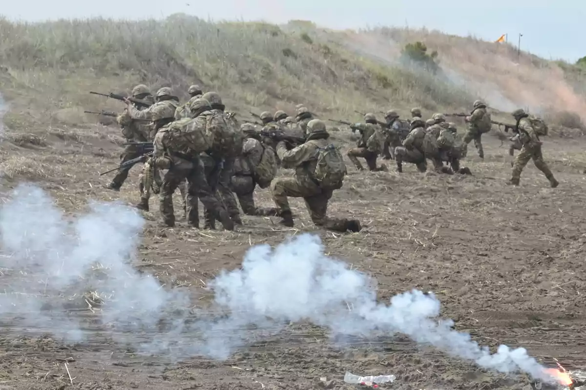 Soldados en uniforme camuflado participan en un ejercicio militar en un campo abierto con humo en el suelo.