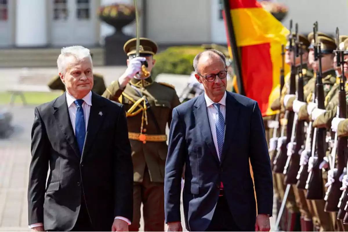 Two men in suits walk in front of a formation of uniformed soldiers with flags at an outdoor official event. Two men in suits walk in front of a formation of uniformed soldiers with flags at an outdoor official event.