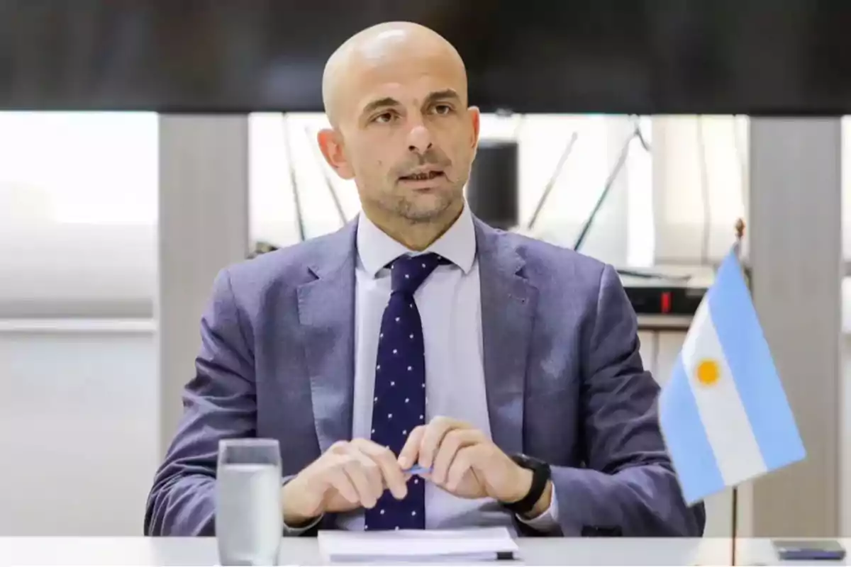A bald man in a suit and tie is sitting at a table with an Argentine flag and a glass of water. A bald man in a suit and tie is sitting at a table with an Argentine flag and a glass of water.