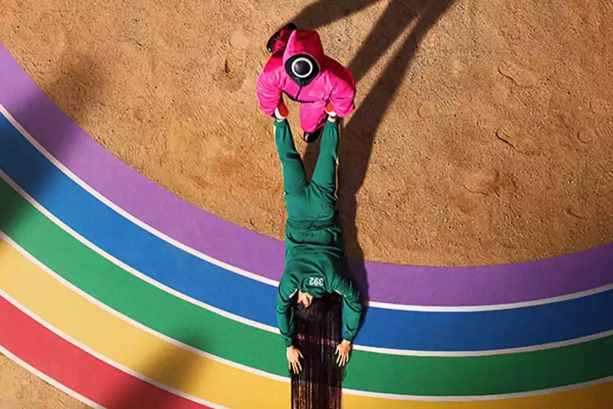 Una persona con un traje verde está acostada en el suelo mientras otra con un traje rosa se encuentra de pie sobre una pista de colores en un entorno arenoso.