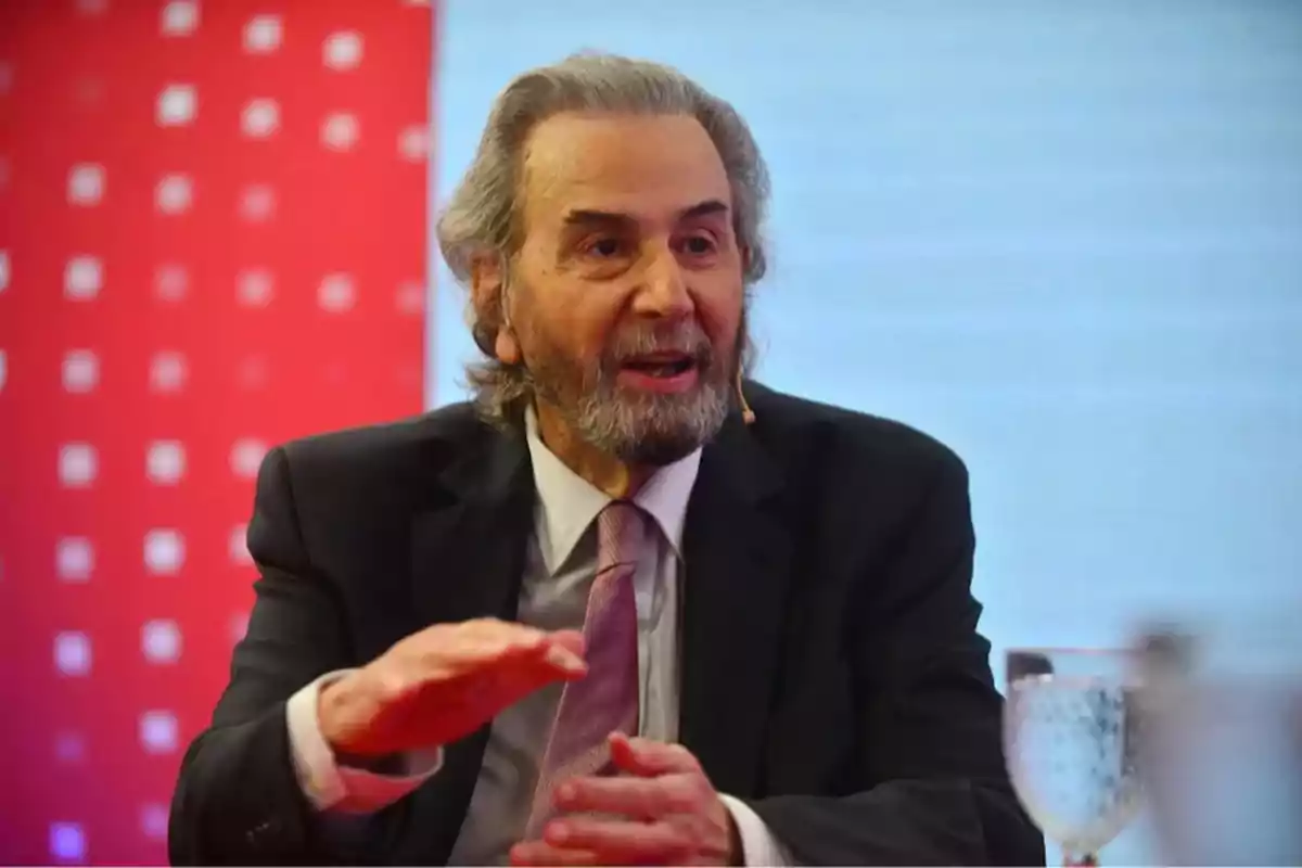 Hombre de cabello canoso y barba hablando en un evento con fondo rojo y azul, viste traje oscuro y corbata clara, tiene un micrófono en la mejilla y un vaso de agua frente a él. Hombre de cabello canoso y barba hablando en un evento con fondo rojo y azul, viste traje oscuro y corbata clara, tiene un micrófono en la mejilla y un vaso de agua frente a él.