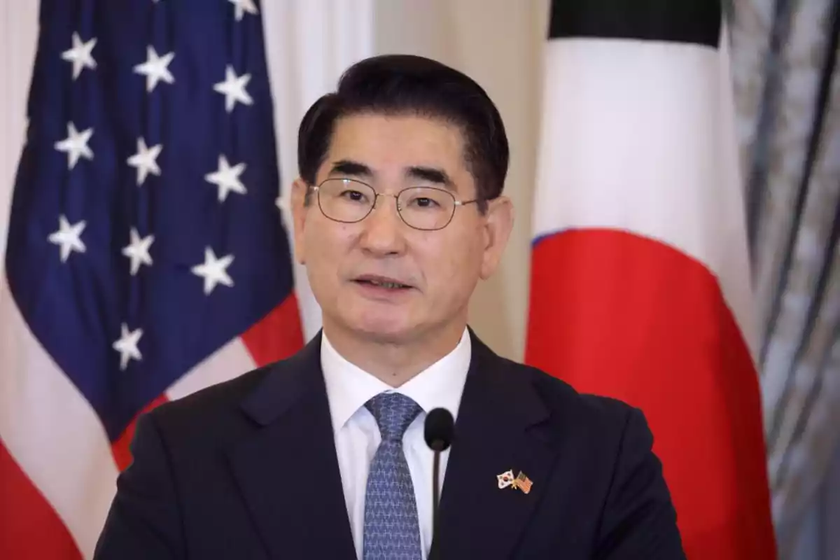 Un hombre con gafas y traje oscuro habla frente a micrófono con las banderas de Estados Unidos y Corea del Sur de fondo.