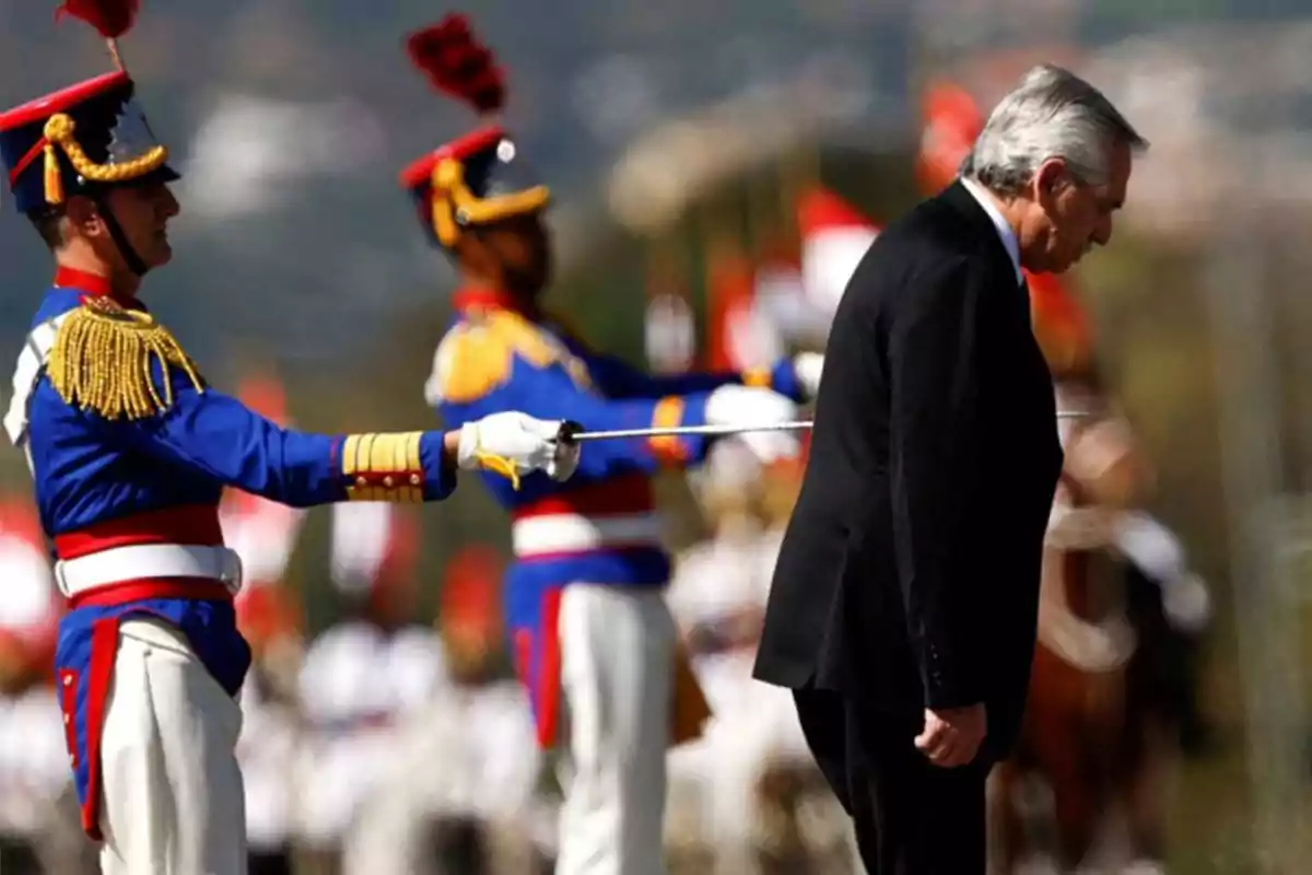 Un hombre de traje negro inclina la cabeza mientras dos soldados con uniformes coloridos y espadas están de pie detrás de él.