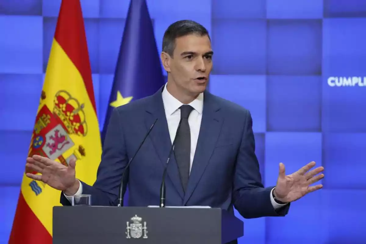Hombre con traje azul hablando en un podio frente a las banderas de España y la Unión Europea sobre un fondo azul Hombre con traje azul hablando en un podio frente a las banderas de España y la Unión Europea sobre un fondo azul