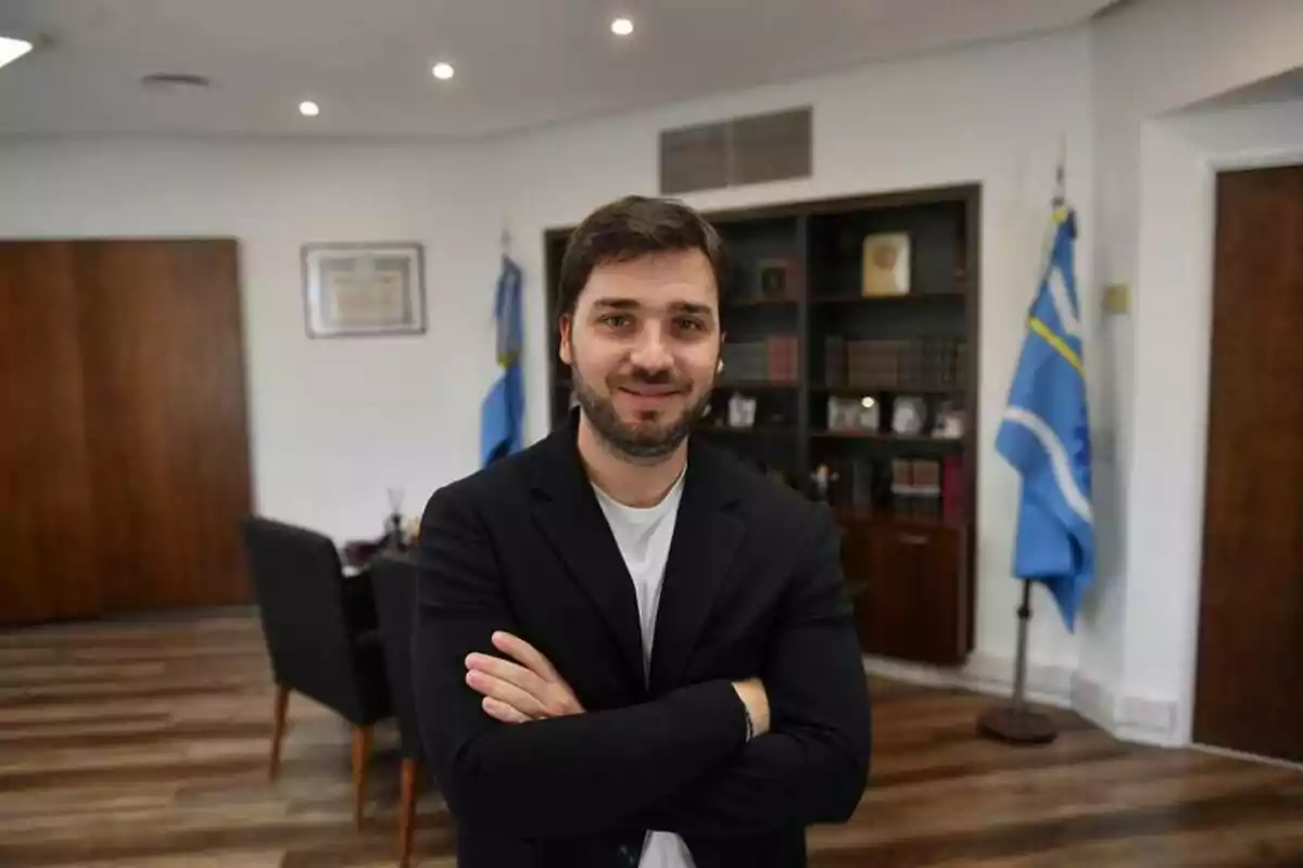 Un hombre de pie con los brazos cruzados en una oficina con banderas y estanterías al fondo.