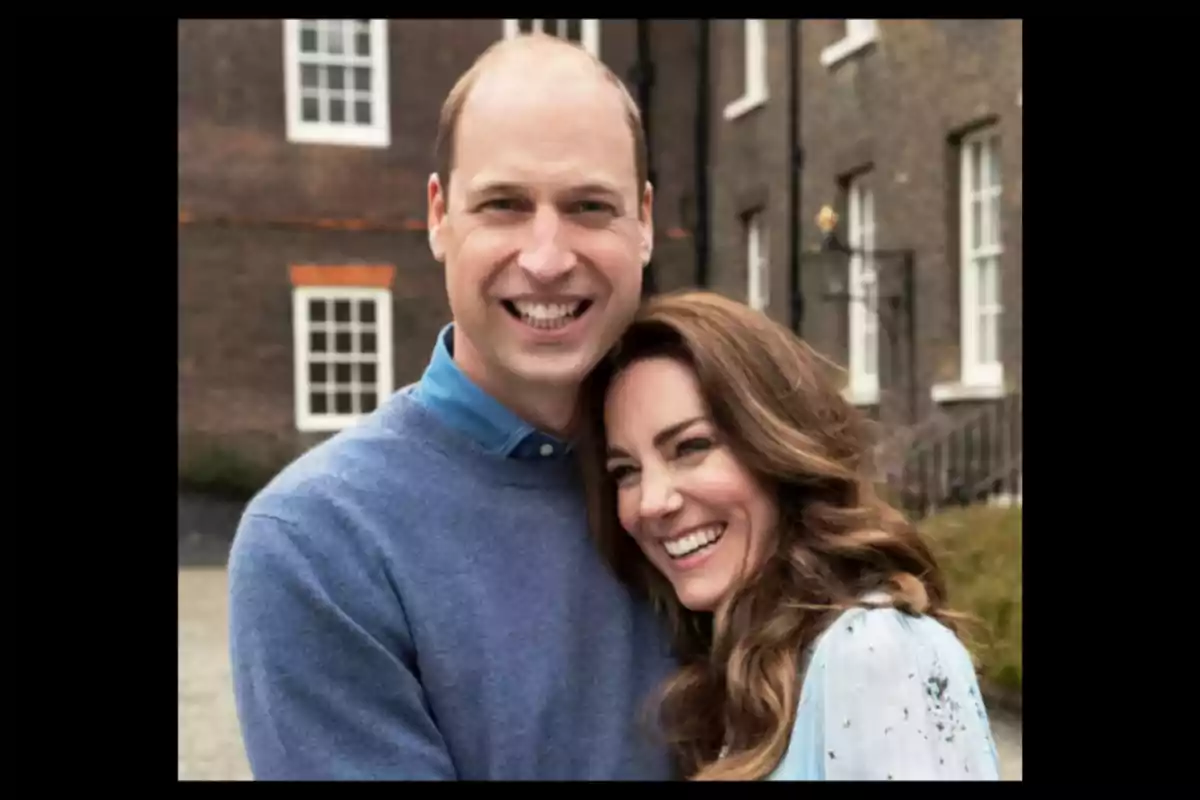 Una pareja sonriente se abraza frente a un edificio de ladrillos con ventanas blancas.