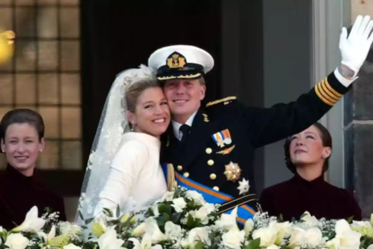 Una pareja sonriente en traje de boda, con el hombre saludando, rodeados de flores blancas y dos personas al fondo.