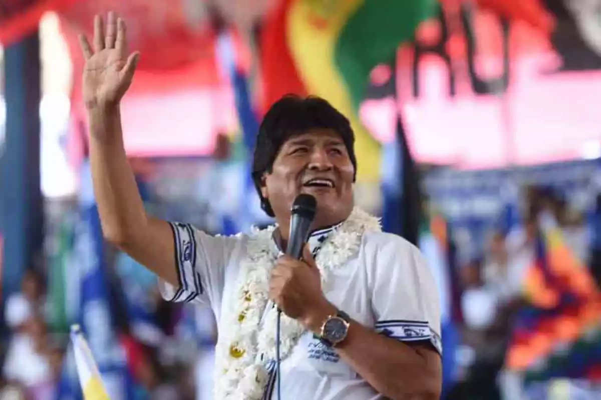 Una persona sonriente con una guirnalda de flores alrededor del cuello sostiene un micrófono y levanta la mano en un evento con banderas de fondo. Una persona sonriente con una guirnalda de flores alrededor del cuello sostiene un micrófono y levanta la mano en un evento con banderas de fondo.