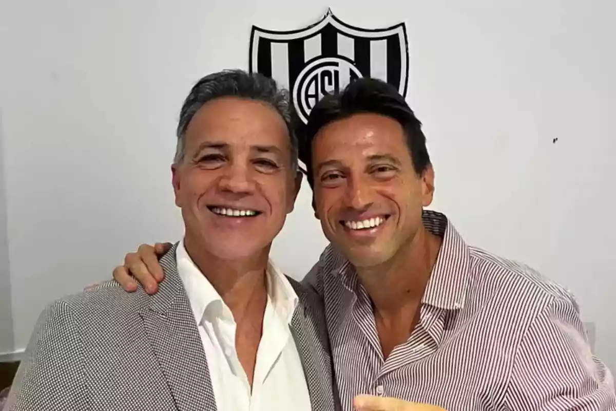 Dos personas sonrientes posan juntas frente a un escudo en la pared. Dos personas sonrientes posan juntas frente a un escudo en la pared.