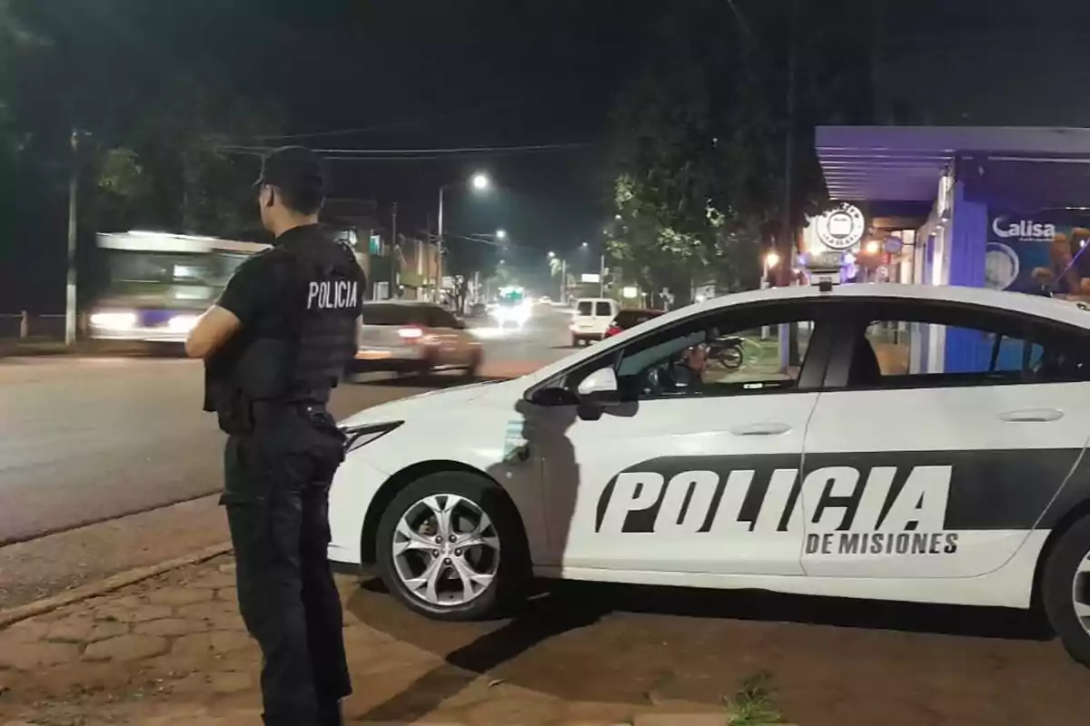 Un policía de pie junto a un coche patrulla de la Policía de Misiones en una calle iluminada por la noche.