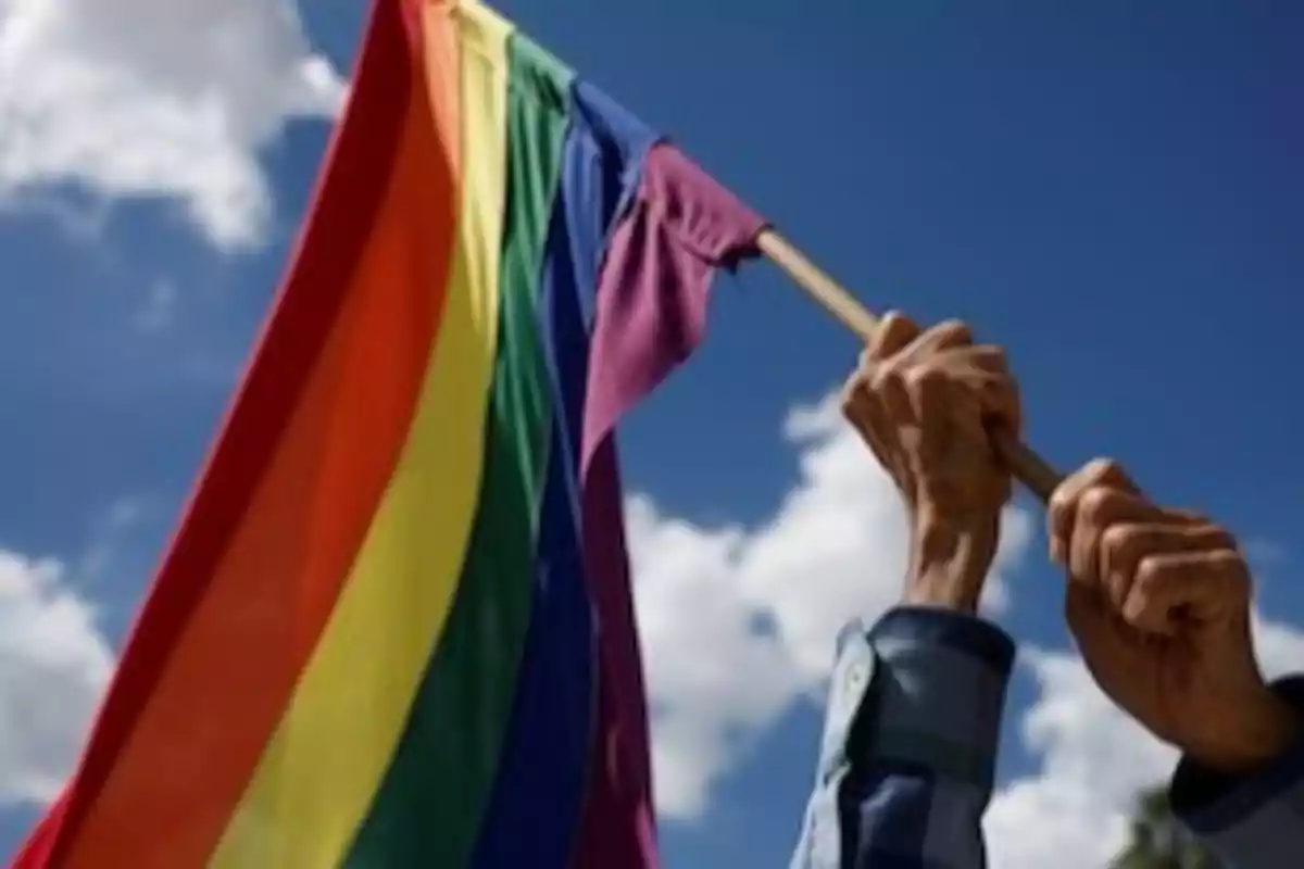 Manos sosteniendo una bandera arcoíris ondeando contra un cielo azul. Manos sosteniendo una bandera arcoíris ondeando contra un cielo azul.