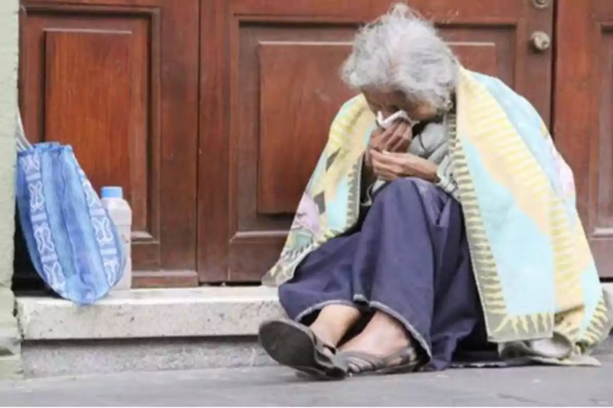 Una persona mayor está sentada en el suelo frente a una puerta de madera, cubierta con una manta y sosteniendo un pañuelo, con una bolsa azul y una botella de agua a su lado. Una persona mayor está sentada en el suelo frente a una puerta de madera, cubierta con una manta y sosteniendo un pañuelo, con una bolsa azul y una botella de agua a su lado.