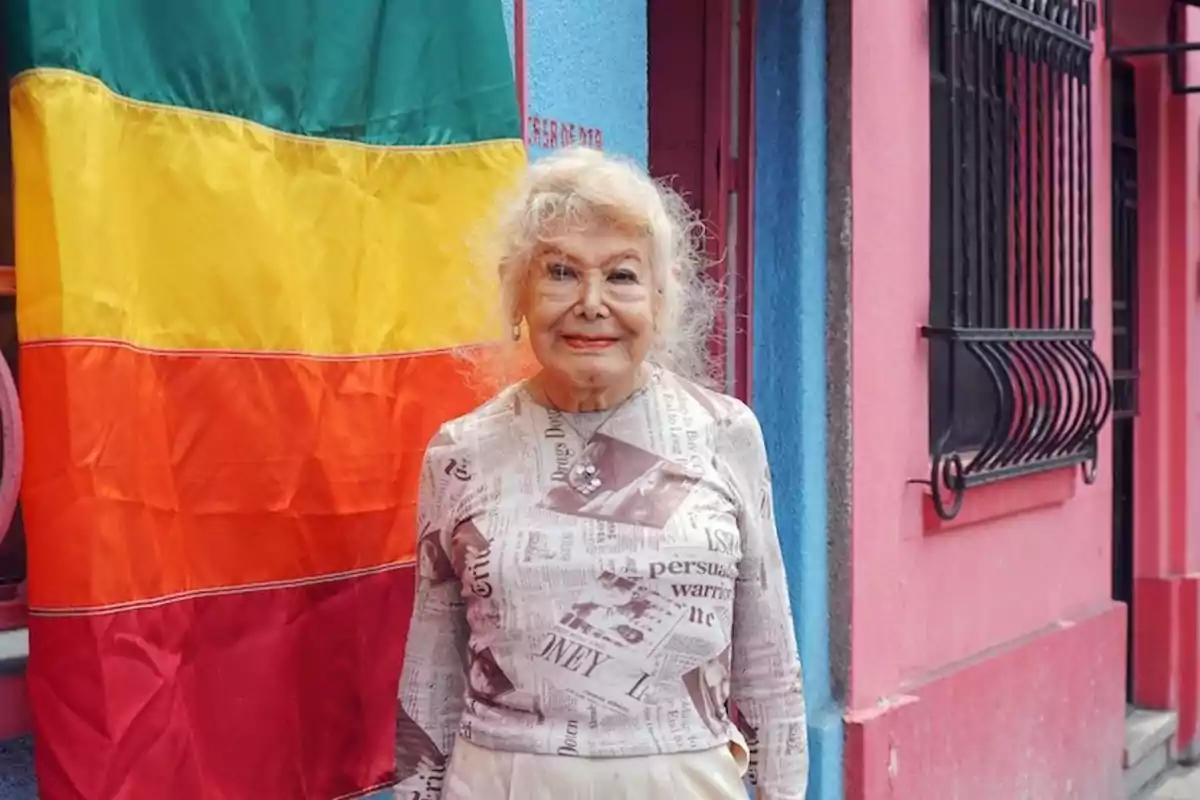 Una persona mayor sonríe frente a una bandera de colores verde, amarillo, naranja y rojo, con una pared de fondo en tonos azul y rosa.