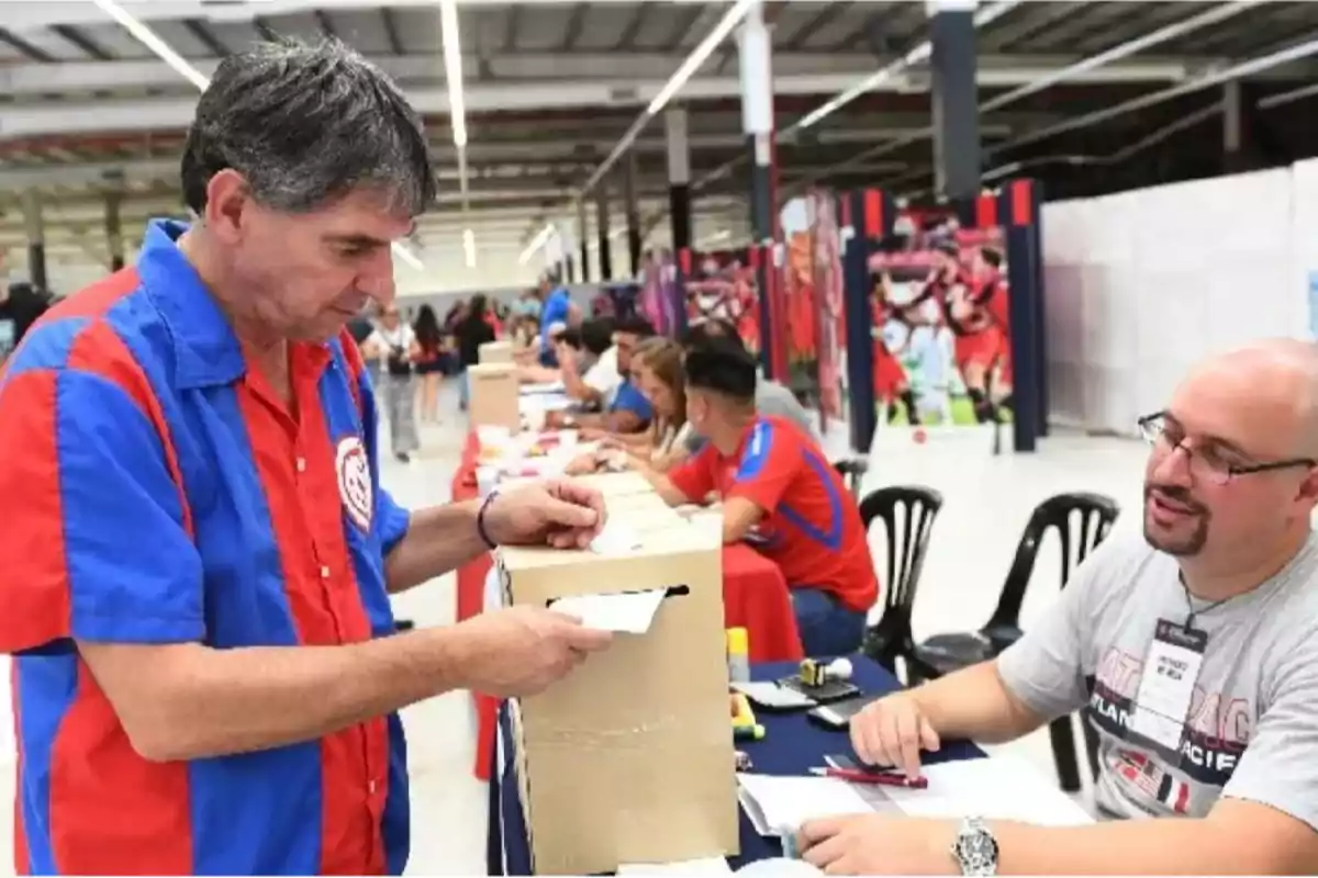 Un hombre con camisa azul y roja deposita un papel en una caja mientras otra persona lo observa en un entorno con varias mesas y personas. Un hombre con camisa azul y roja deposita un papel en una caja mientras otra persona lo observa en un entorno con varias mesas y personas.