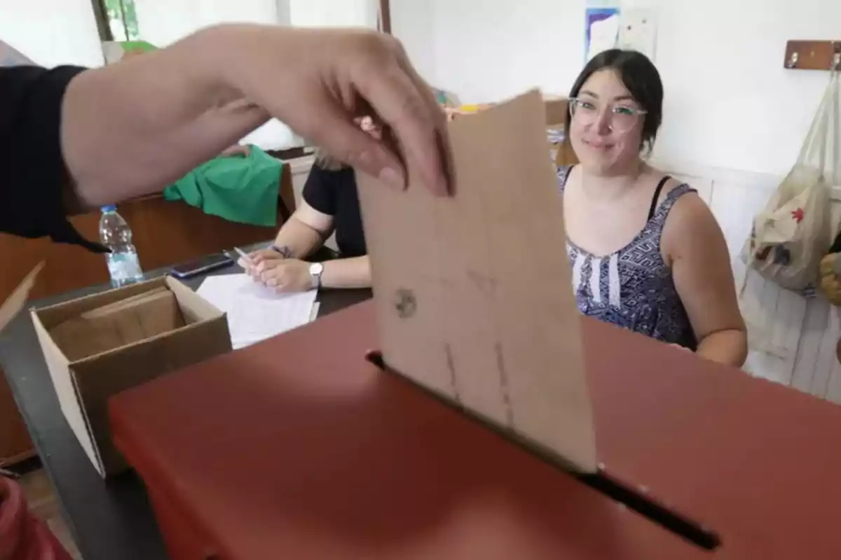 Una persona depositando un sobre en una urna mientras otra persona observa en un entorno de votación.