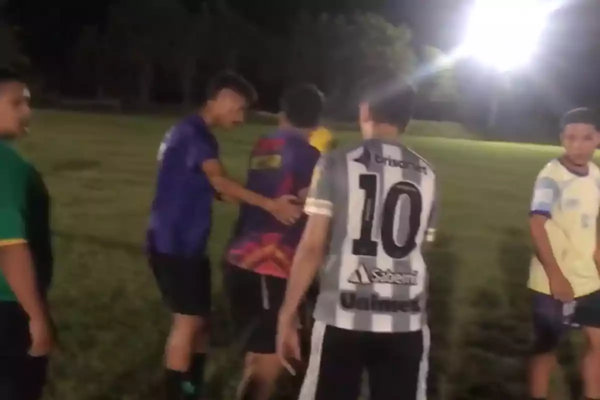 Un grupo de jóvenes en un campo de fútbol iluminado por focos, algunos llevan camisetas deportivas y parecen estar interactuando después de un partido. Un grupo de jóvenes en un campo de fútbol iluminado por focos, algunos llevan camisetas deportivas y parecen estar interactuando después de un partido.