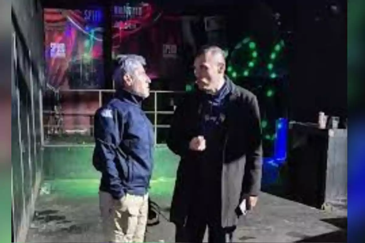 Dos hombres conversan de pie en el interior de un lugar oscuro con luces de colores al fondo. Dos hombres conversan de pie en el interior de un lugar oscuro con luces de colores al fondo.