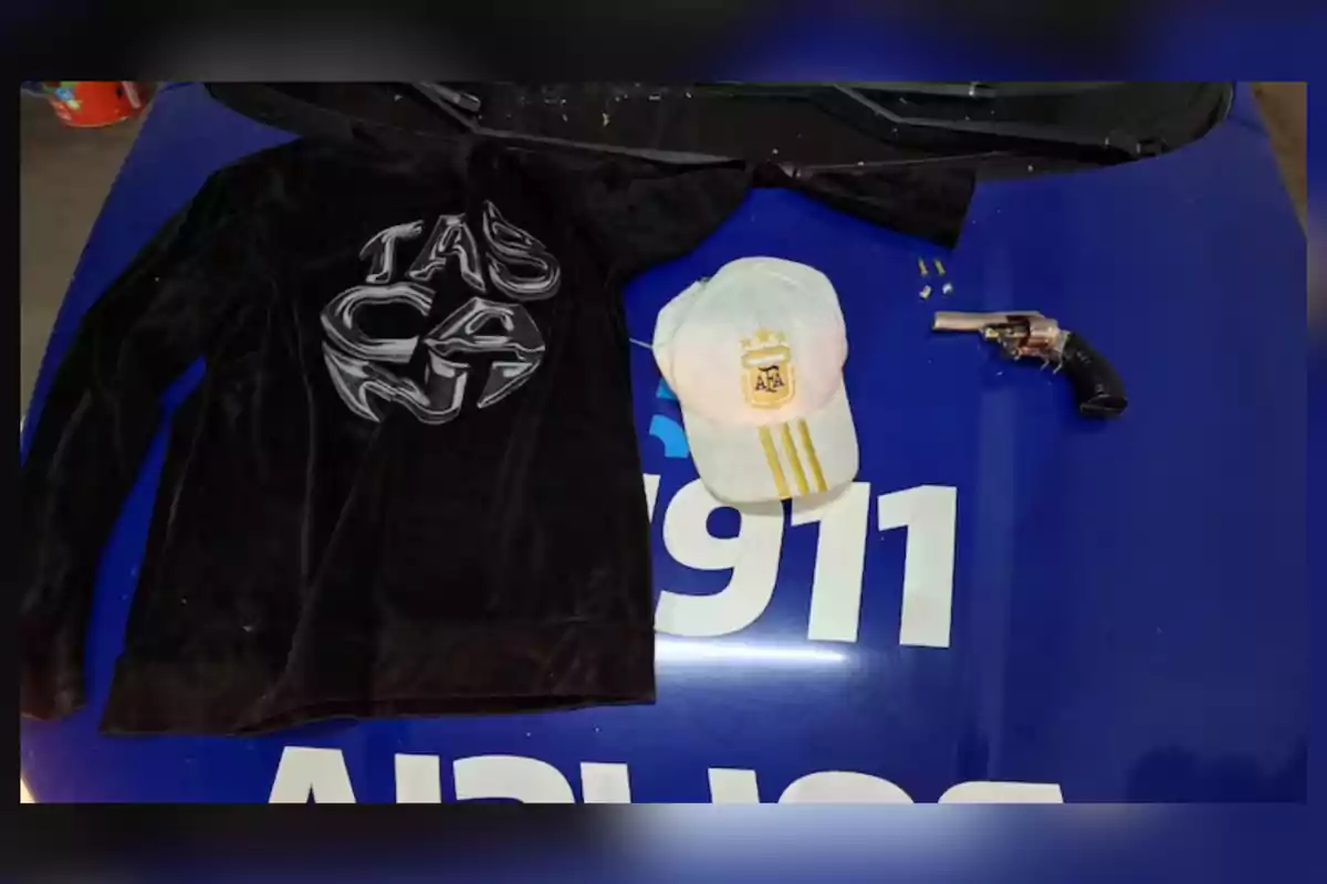 Sobre el cofre de un auto azul con el número 911 se observa una sudadera negra, una gorra blanca con el escudo de la AFA, un revólver y varias balas. Sobre el cofre de un auto azul con el número 911 se observa una sudadera negra, una gorra blanca con el escudo de la AFA, un revólver y varias balas.