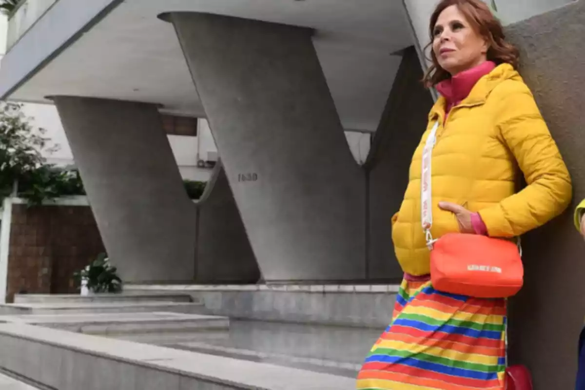 Mujer con chaqueta amarilla y bolso naranja posando frente a un edificio moderno con columnas en forma de V.