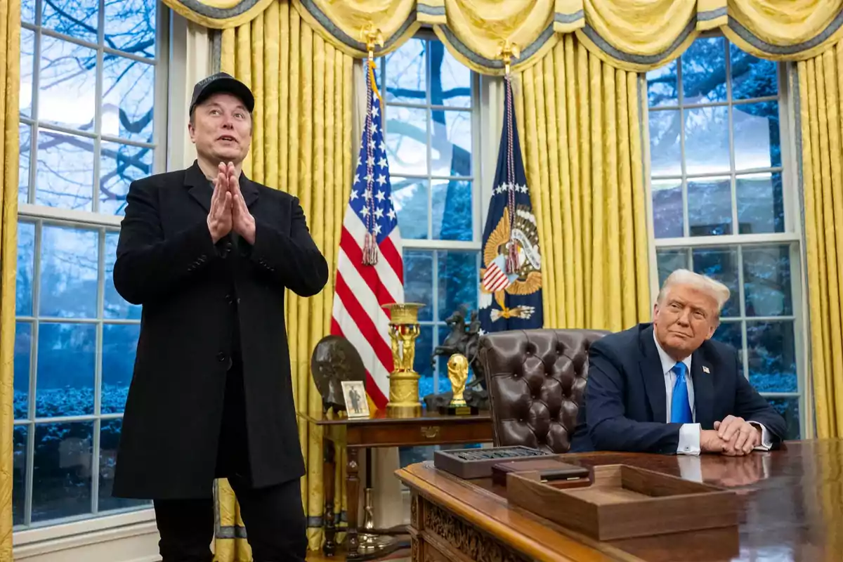 Two people in an office with golden curtains and American flags, one standing with hands together and the other sitting at a desk. Two people in an office with golden curtains and American flags, one standing with hands together and the other sitting at a desk.