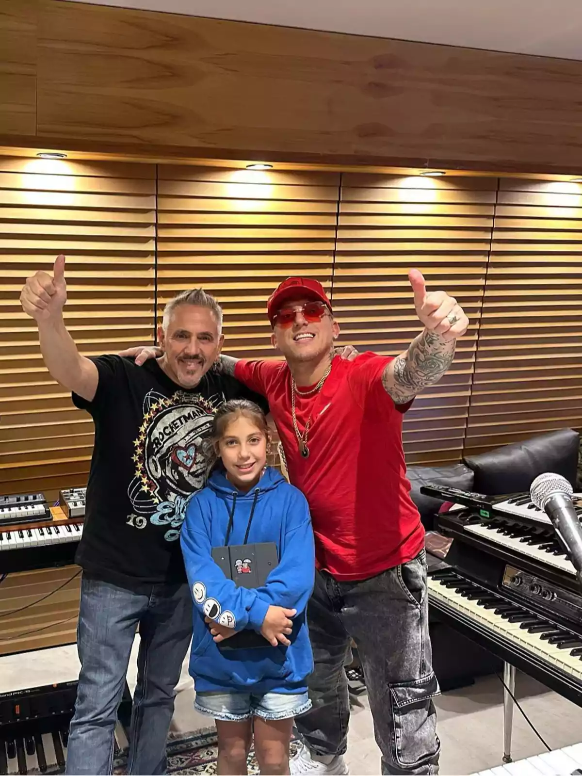Three people smile as they pose in a recording studio; two adults give a thumbs up while a girl in a blue sweatjersey holds a tablet in front of several keyboards and synthesizers. Three people smile as they pose in a recording studio; two adults give a thumbs up while a girl in a blue sweatjersey holds a tablet in front of several keyboards and synthesizers.