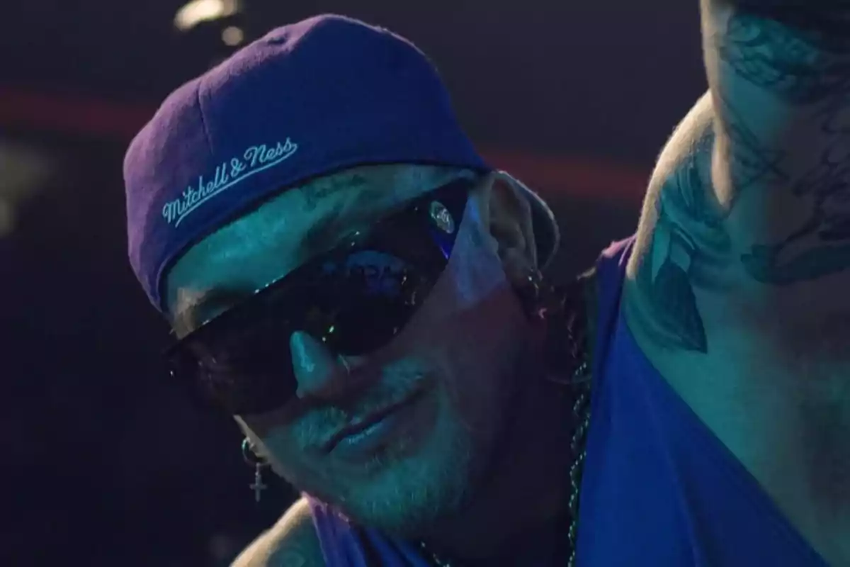 Hombre con gorra morada, gafas oscuras y tatuajes en el cuello, posando en un ambiente con luz tenue