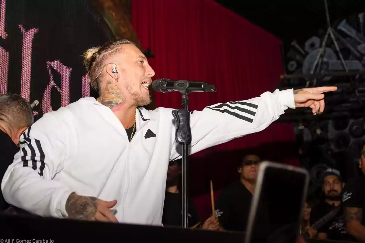 Cantante con chaqueta blanca y tatuaje en el cuello interpretando una canción en el escenario mientras señala hacia el público Cantante con chaqueta blanca y tatuaje en el cuello interpretando una canción en el escenario mientras señala hacia el público