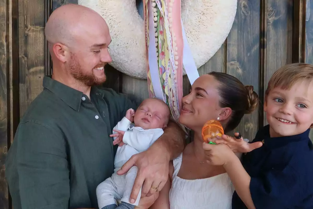 Una familia sonriente posa frente a una puerta de madera, el padre sostiene a un bebé mientras la madre y un niño pequeño miran con alegría. Una familia sonriente posa frente a una puerta de madera, el padre sostiene a un bebé mientras la madre y un niño pequeño miran con alegría.