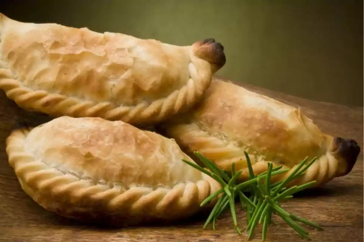Three golden empanadas on a wooden board with a sprig of rosemary in front. Three golden empanadas on a wooden board with a sprig of rosemary in front.